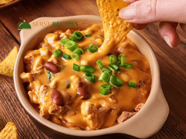 Close-up of a hand dipping a tortilla chip into a thick, cheesy buffalo chicken dip garnished with green onions.