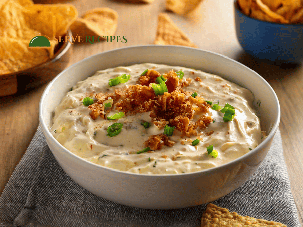 Smooth buffalo chicken dip in a white ceramic bowl, topped with crumbled bacon and served with chips.