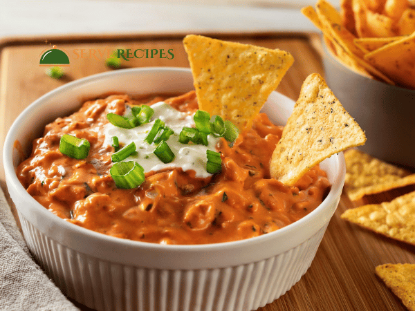 A traditional buffalo chicken dip in a ceramic dish, garnished with sour cream and chives, served with a side of chips.