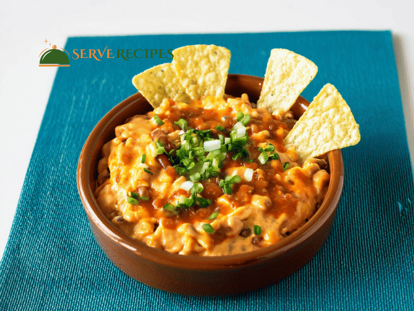 Creamy Buffalo Chicken Queso Dip garnished with green onions, served with blue corn tortilla chips, celery, and carrot sticks on a wooden table.